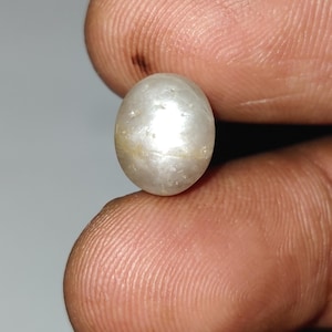 May include: A close-up of a lustrous, oval-shaped pearl. The pearl has a creamy white color with subtle variations and a smooth, reflective surface. The pearl is held between two fingers.