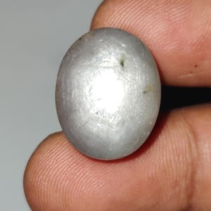 May include: A smooth, oval-shaped, gray gemstone held between fingers. The stone has a slightly pearlescent sheen and visible surface imperfections. The gem is a neutral color, suitable for jewelry or collection.