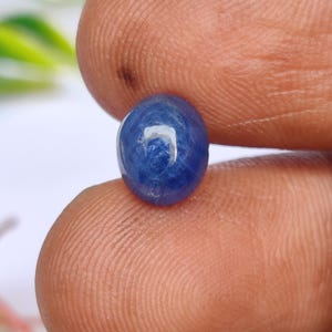 May include: A small, oval-shaped blue sapphire gemstone with a smooth, polished cabochon surface. The stone displays a deep, saturated blue colour with a visible light reflection on its rounded top, showcasing a translucent and polished appearance.
