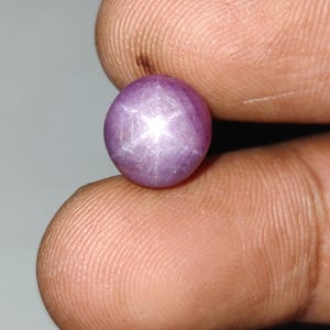 May include: A round, cabochon-cut star ruby gemstone with a translucent, purplish-pink hue. The stone displays a distinct six-rayed star effect, known as asterism, which appears as a bright white light reflection centered on the polished surface.