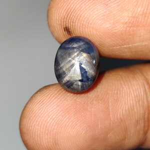 Op de afbeelding: Een ovaalvormige cabochon edelsteen met een diepblauwe kleur en een duidelijk zesstralig sterpatroon op het gepolijste oppervlak. De steen vertoont interne insluitingen en lichtreflecties die een glinsterend effect over het blauw creëren.