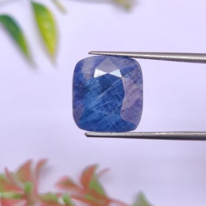 May include: A faceted, cushion-cut sapphire gemstone held by tweezers. The sapphire displays a deep blue hue with lighter internal facets. The gemstone is rectangular with rounded corners, set against a soft, blurred background.