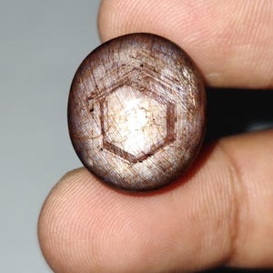 May include: A round, cabochon-cut star sapphire gemstone with a dark brownish-grey hue. The surface displays a distinct, centred six-rayed star pattern formed by light reflection, with visible internal fibrous inclusions and a hexagonal growth structure.