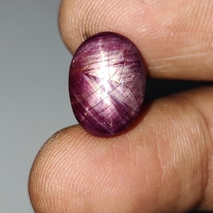 Könnte beinhalten: Ein ovaler Sternrubin-Edelstein im Cabochon-Schliff mit einem tiefen purpurroten Farbton. Der Stein weist einen ausgeprägten sechsstrahligen Sterneffekt auf, der als Asterismus bekannt ist und auf seiner polierten Oberfläche sichtbar ist.