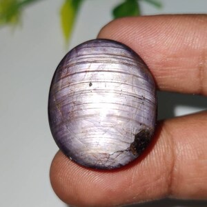 Puede incluir: Una gran piedra preciosa ovalada de color púrpura con un brillo plateado y estrías visibles. La piedra preciosa se sostiene en una mano.