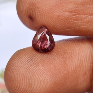 May include: A faceted, pear-shaped gemstone with a deep reddish-pink hue. The gem is held between fingers, showcasing its polished surfaces and internal reflections. The stone's cut highlights its clarity and brilliance, making it a striking piece of jewelry.