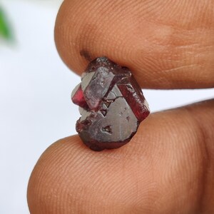 Puede incluir: Una pequeña piedra preciosa de granate en bruto de color rojo intenso con una estructura cristalina natural multifacética. La piedra muestra un tono burdeos oscuro y translúcido con facetas geométricas visibles y bordes afilados.