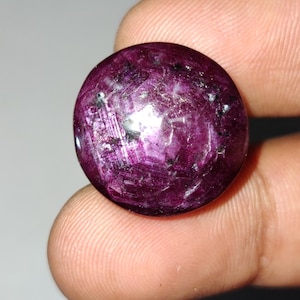 May include: A round, polished gemstone with a deep magenta hue, exhibiting internal light reflections. The gem has a smooth, rounded surface and appears to be a ruby. The stone is held between fingers, showcasing its size and color.