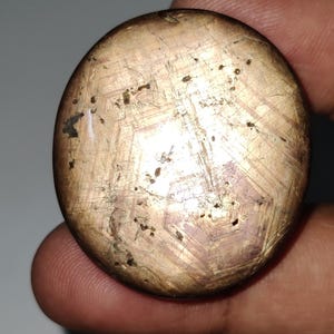 Può includere: Una pietra preziosa rotonda tagliata a cabochon con un motivo a forma di stella. La pietra è di colore marrone chiaro con un tocco di rosa e ha una finitura scintillante e iridescente.