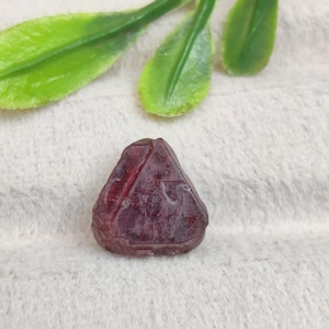 May include: A small, triangular-shaped deep red gemstone with a faceted surface and visible internal inclusions. The stone has a polished appearance with distinct geometric lines carved into its top face, showcasing a dark burgundy hue.