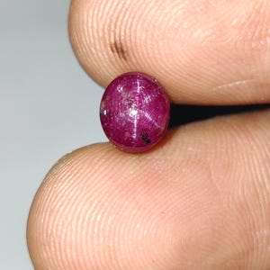 May include: A small, round, deep red ruby gemstone. The polished surface of the gem reflects light, showcasing internal structures. The ruby is held between fingers, highlighting its size and color. This gemstone is suitable for jewelry making.