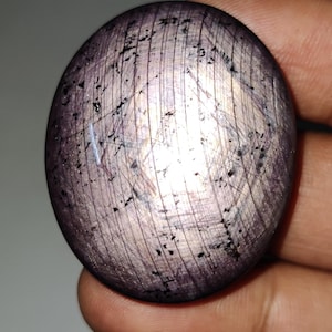 Op de afbeelding: Een ronde, cabochon geslepen edelsteen met een zilverachtig-paarse tint en zwarte insluitsels. De steen heeft een glad, gepolijst oppervlak en wordt in een hand gehouden.