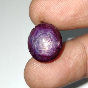 May include: A round, purple gemstone with a star-shaped pattern inside. The gemstone is held in a person's hand.
