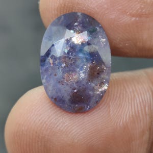 Puede incluir: Una gema ovalada y facetada con una mezcla de tonos azules, morados y marrones. La gema tiene una calidad brillante e iridiscente, con inclusiones internas que se suman a su apariencia única. La piedra se sostiene entre dos dedos.