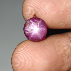May include: A round, polished, purplish-red gemstone with a star-like effect. The gem is held between two fingers, showcasing its color and internal structure. The stone has a smooth, reflective surface.