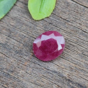 Könnte beinhalten: Ein facettierter, ovaler Rubin-Edelstein mit tiefroter Farbe. Der Edelstein liegt auf einer rustikalen Holzoberfläche, mit grünen Blättern im Hintergrund. Der Schliff des Rubins erzeugt mehrere reflektierende Flächen.