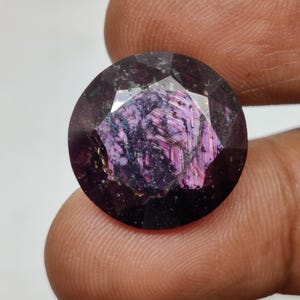 Può includere: Una pietra preziosa rotonda e sfaccettata con una tavolozza di colori viola intenso e magenta scuro. La pietra mostra una struttura interna complessa con inclusioni visibili, venature scure e una superficie lucida che riflette la luce.