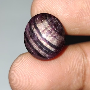 May include: A round, polished gemstone with a mix of purple and silver-white bands. The stone has a smooth, reflective surface and appears to be held between fingers. The gemstone's color and pattern create a unique, eye-catching appearance.
