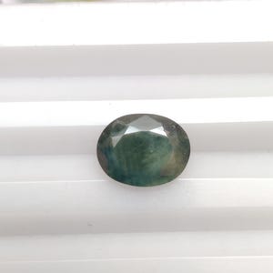 May include: A round, faceted gemstone with a dark green and blue colour. The gemstone has a slightly cloudy appearance.