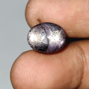 Puede incluir: Primer plano de una gema ovalada pulida. La gema tiene una mezcla de colores, incluyendo púrpura, plata y negro, con un efecto de estrella visible en su superficie. La piedra se sostiene entre dos dedos.