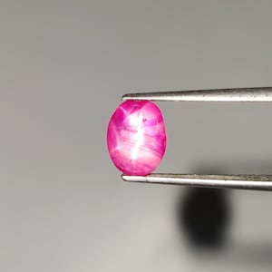 Könnte beinhalten: Ein leuchtend pinkfarbener Sternrubin in ovaler Form mit einem ausgeprägten sechsstrahligen Sterneffekt auf seiner polierten, durchscheinenden Oberfläche. Der Edelstein wird von einer Metallpinzette gehalten und zeigt seinen tiefen Magentaton.