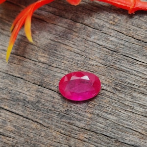 May include: A vibrant pink oval-shaped gemstone with a faceted cut, resting on a rustic wooden surface.