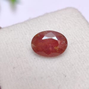 May include: A faceted, oval-shaped gemstone with a reddish-brown hue. The gem is set against a white background, showcasing its polished surfaces and internal clarity. The gemstone's cut allows light to reflect, creating a sparkling effect.