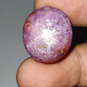 May include: Oval-shaped, purplish-pink gemstone with a star-like pattern. The gem has a polished surface with visible inclusions and a subtle color variation. The stone is held between fingers, showcasing its size and shape.