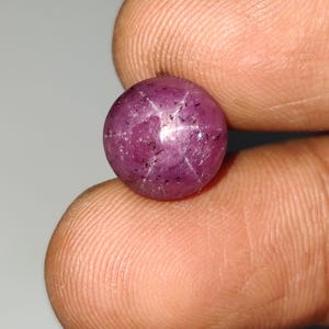 Pode incluir: Uma pedra preciosa de rubi estrela de corte cabochão redondo com um tom magenta-rosa profundo. A pedra apresenta um efeito de estrela de seis raios visível na sua superfície polida e abobadada, com inclusões escuras naturais dispersas.