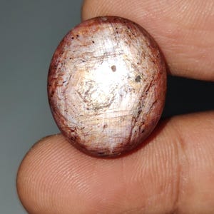 Könnte beinhalten: Ein oval geschliffener Edelstein mit einer Mischung aus rötlich-braunen, weißen und goldenen Farbtönen. Die Oberfläche weist eine strukturierte Optik mit feinen Mustern und winzigen dunklen Einschlüssen auf. Der Edelstein wird zwischen Fingern gehalten.