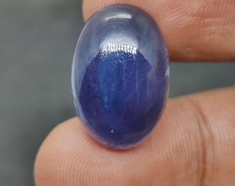 May include: A polished, oval-shaped, deep blue gemstone. The gem has a smooth, reflective surface, with a slight internal pattern visible. The stone is held between two fingers, showcasing its size and color. The gem appears to be a cabochon cut.