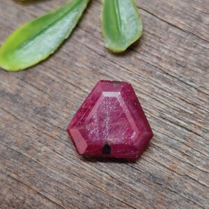 Puede incluir: Una gema de rubí hexagonal y facetada con un tono rojo intenso. La gema tiene una superficie pulida y está colocada sobre un fondo de madera. Se ven hojas verdes en el fondo.