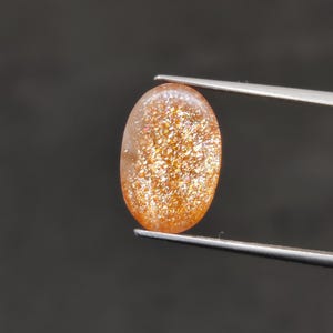 May include: An oval-shaped sunstone gemstone, held by tweezers against a dark background. The stone has a warm, orange-brown color with sparkling inclusions, creating a shimmering effect. The gem is polished and smooth.