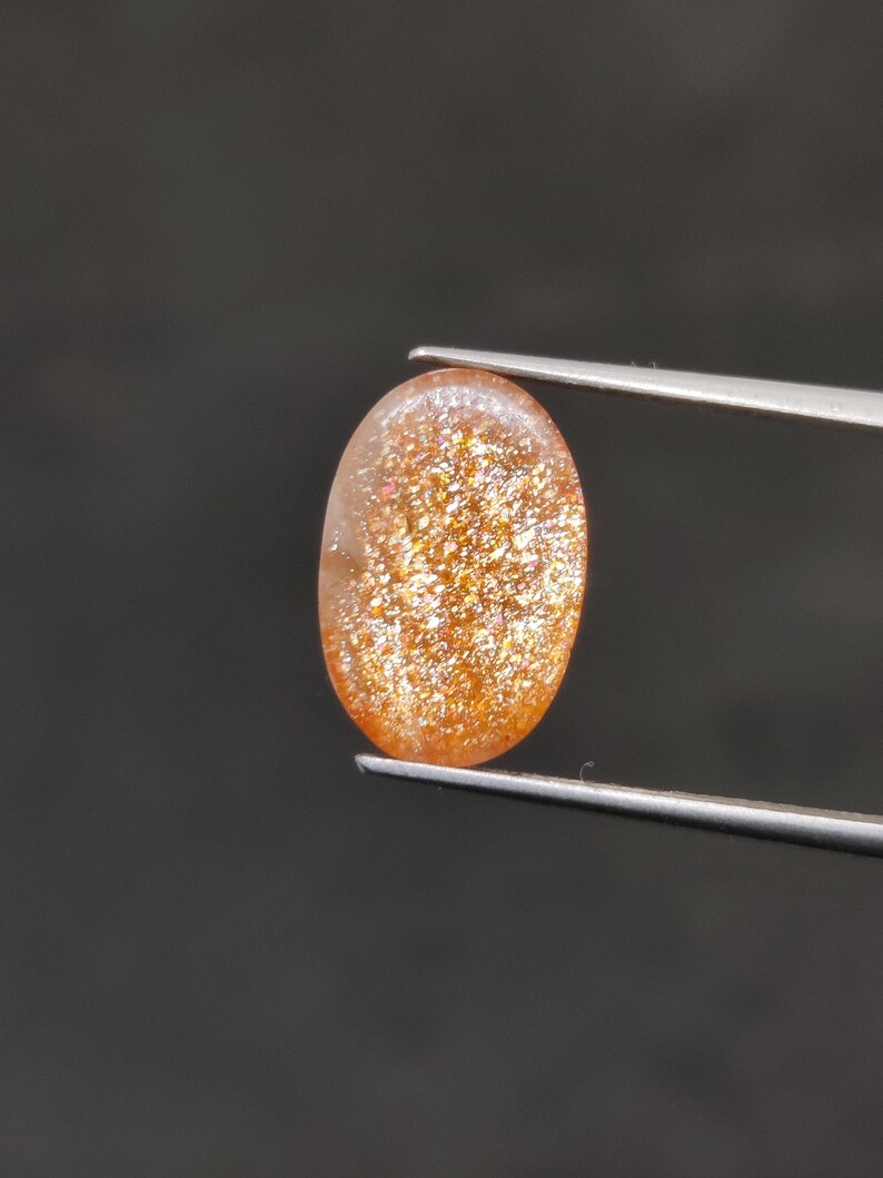 May include: An oval-shaped sunstone gemstone, held by tweezers against a dark background. The stone has a warm, orange-brown color with sparkling inclusions, creating a shimmering effect. The gem is polished and smooth.