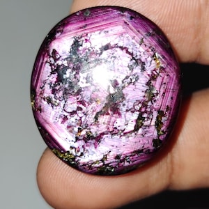 Pode incluir: Uma gema redonda e polida com um tom magenta profundo. A pedra apresenta um padrão em forma de estrela e inclusões internas pretas e douradas. A superfície reflete a luz, criando um efeito brilhante. A gema é segurada entre os dedos.