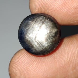 May include: A dark, round gemstone with a star-like pattern. The gem has a polished surface with a mix of gray and blue hues. The star effect is created by intersecting lines across the surface, creating a six-pointed star.