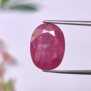 May include: A faceted, oval ruby gemstone held by tweezers. The ruby is a deep, saturated red colour with visible internal inclusions. The gemstone is displayed against a soft, blurred background.