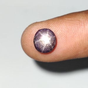 Puede incluir: Una gema redonda y pulida, de color púrpura, con un patrón de estrella de seis rayos en su superficie. La gema se sostiene entre dos dedos, mostrando su color y cualidades reflectantes. La piedra preciosa es de color púrpura oscuro.
