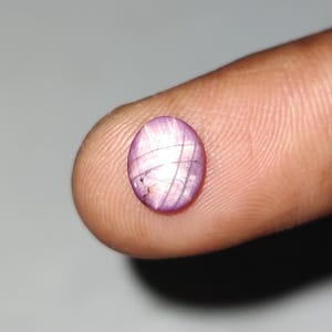 May include: Oval-shaped, purplish-pink gemstone with a star-like effect. The gem has a polished surface with intersecting lines creating a star pattern. The stone is held between two fingers against a neutral background. Gemstone for jewelry.