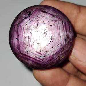 Può includere: Una gemma rotonda e lucidata di colore magenta intenso. La pietra presenta un motivo a stella al centro, con inclusioni visibili e un design esagonale. La gemma è tenuta in una mano.