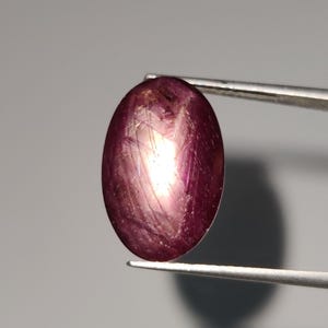 Könnte beinhalten: Ein ovaler Edelstein in tiefroter Farbe mit einem sternförmigen Muster in der Mitte. Der Edelstein wird von einer Pinzette gehalten.