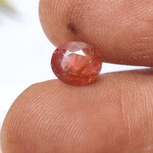 Puede incluir: Una gema redonda y facetada, con un cálido tono rojo-anaranjado. La gema se sostiene entre dos dedos, mostrando su superficie pulida y claridad interna. La piedra preciosa es una piedra suelta, adecuada para la creación de joyas.