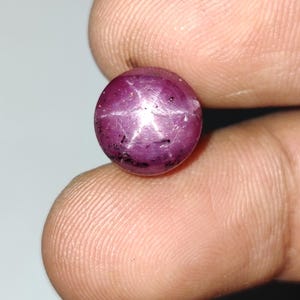 Op de afbeelding: Een ronde, in cabochon geslepen ster-robijn edelsteen met een diepe magenta-rode kleur en een duidelijk zesstralig stereffect, bekend als asterisme, zichtbaar op het gepolijste oppervlak, samen met kleine donkere insluitingen in de steen.