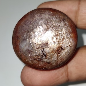 Könnte beinhalten: Ein runder, polierter Edelstein mit einer Mischung aus Braun-, Rotbraun- und Silbertönen. Die Oberfläche hat eine schimmernde, reflektierende Qualität mit sichtbaren Einschlüssen und einer leicht unebenen Textur. Der Stein wird von Fingern gehalten.