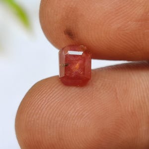 Op de afbeelding: Een kleine, rechthoekig geslepen, rood-oranje edelsteen. De edelsteen wordt tussen twee vingers gehouden, waardoor de facetten en de interne structuur zichtbaar worden. De steen heeft een warme kleur en een gepolijste uitstraling.