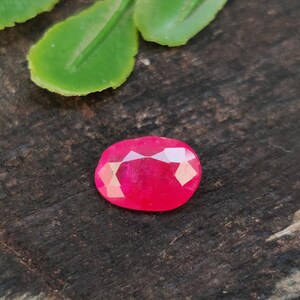 May include: A faceted, oval-shaped, vibrant red gemstone. The gem is set against a dark, textured wooden surface, with a green leaf in the background. The gemstone's cut creates multiple reflective surfaces, enhancing its brilliance.