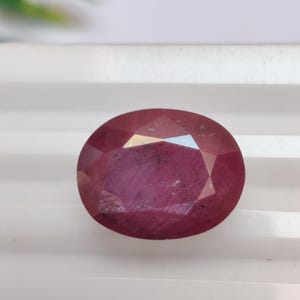 May include: A faceted, oval-shaped ruby gemstone. The gem is a deep red colour with internal inclusions. The stone is set against a white background, with a hint of green foliage in the upper left corner.