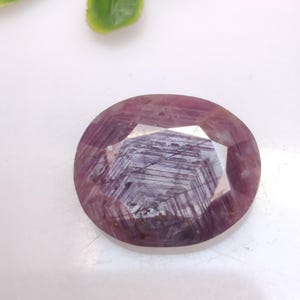 Puede incluir: Una gema facetada, de forma ovalada, con un tono rosa intenso. La gema tiene una estructura interna compleja con estrías visibles y una superficie pulida. El fondo es blanco, con algunas hojas verdes en la esquina superior izquierda. Mide unos 2.5 cm.