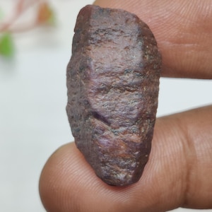 May include: A rough, uncut ruby gemstone with a deep reddish-brown hue. The natural stone has a textured surface and is held between two fingers. The ruby is a raw, unpolished specimen, showcasing its natural form.