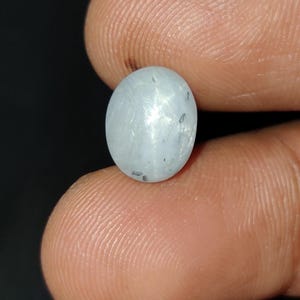 May include: A light blue, oval-shaped gemstone with a star-like effect. The gem is held between fingers against a dark background. The stone has a smooth, polished surface and appears to be translucent.
